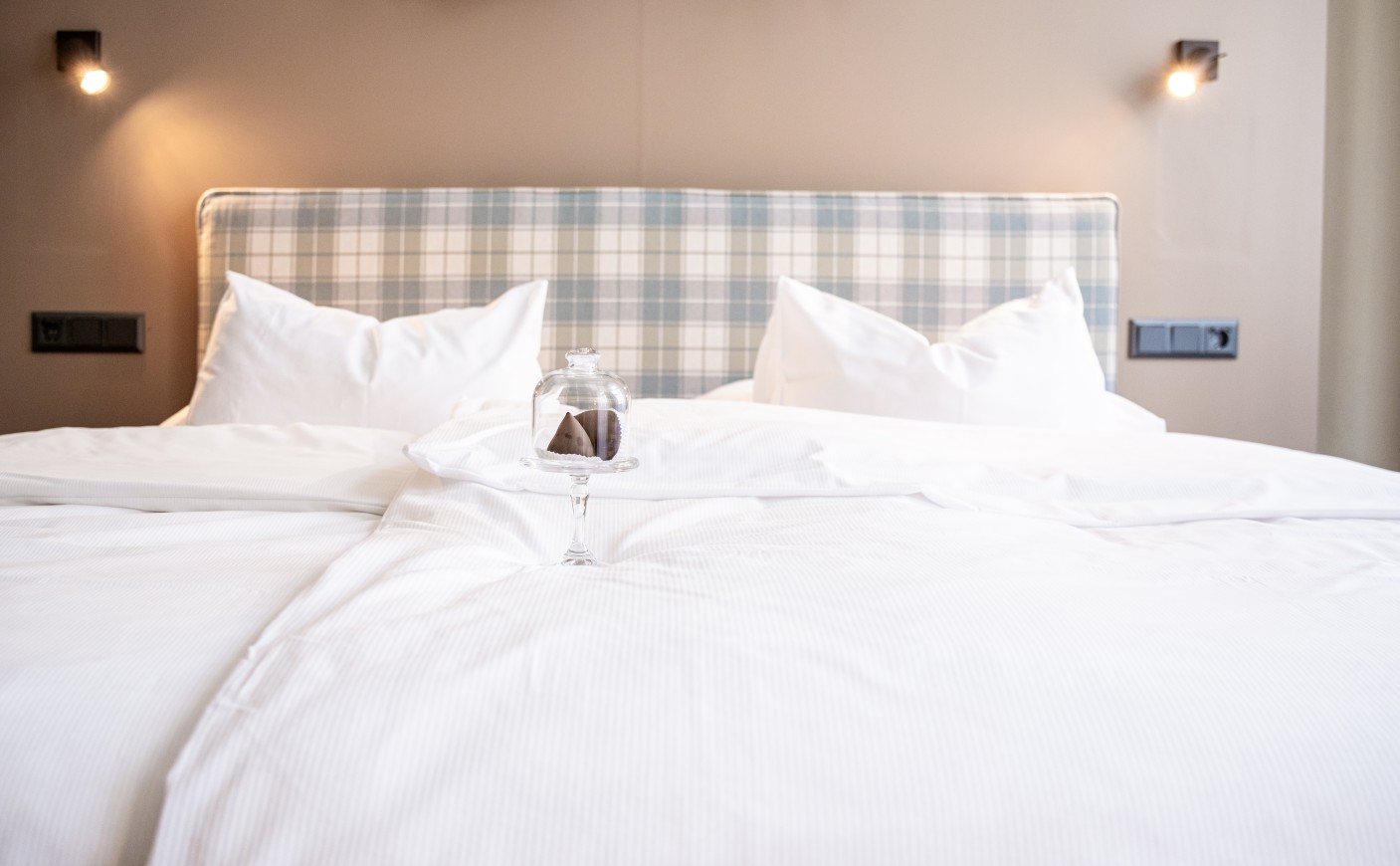 A neatly made bed with white linens and a covered glass dish holding a treat
