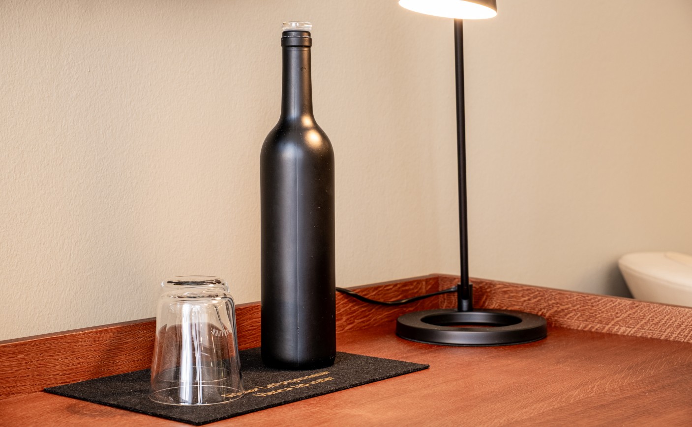 A black bottle and an upside-down glass sit on a wooden desk beside a lamp